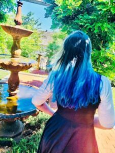 A person with long blue hair is pictured from behind, standing near a water fountain on a sunny day.